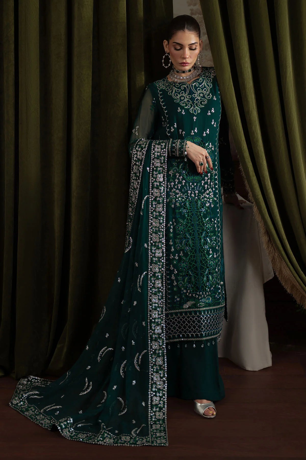Model posing with curtain, showcasing long dupatta and detailed embroidery on Nureh Elanora Style 77 suit
